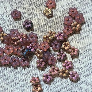 May include: Close-up of a collection of small, flower-shaped beads in shades of purple, bronze, and gold. The beads have a textured, multi-faceted design. The beads are scattered on a textured, off-white surface. Ideal for jewelry making.