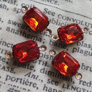 Op de afbeelding: Vier rechthoekige, gefacetteerde, rode glazen juwelen met messingkleurige metalen lussen aan elk uiteinde. De juwelen zijn gezet in een messingkleurig metalen frame. De achtergrond is een vintage stof met vervaagde tekst.