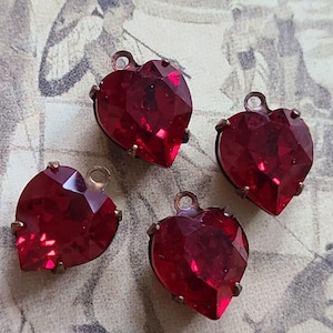 May include: Four faceted, heart-shaped red glass pendants with a bronze-colored metal setting. Each pendant has a small loop at the top for attaching to a chain or other jewelry component. The pendants are displayed on a vintage patterned background.