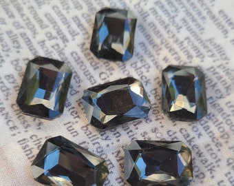 May include: Six faceted, dark gray rectangular and geometric shaped rhinestones. The rhinestones have a reflective surface and are arranged on a patterned fabric background. These could be used for jewelry making or embellishments.