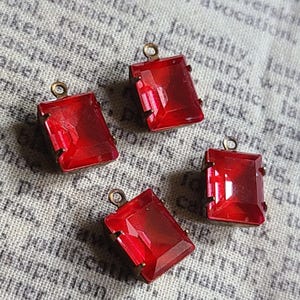 Könnte beinhalten: Vier rote Glas-Anhänger mit goldfarbenen Metallfassungen. Die Anhänger sind rechteckig und haben ein facettierten Design.
