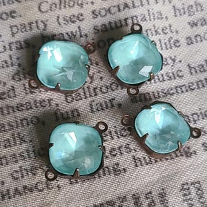 May include: Four square, faceted, light blue glass cabochons with bronze-colored metal settings. Each has a loop for attaching to jewelry or other crafts. The background is a printed fabric.
