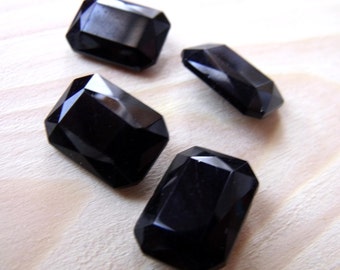 May include: Four black, faceted, rectangular glass gems. The gems are arranged on a light wood surface.