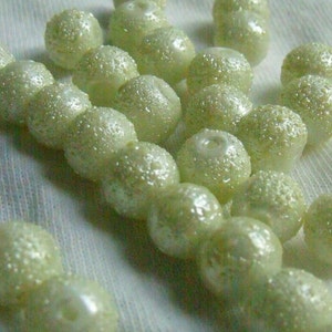 Puede incluir: Una hebra de cuentas de color verde claro con una superficie texturizada. Las cuentas están enhebradas y están sobre una superficie de tela blanca.