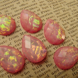 May include: Seven pink teardrop-shaped glass beads with iridescent gold flecks. The beads have a faceted design and are scattered on a brown surface.