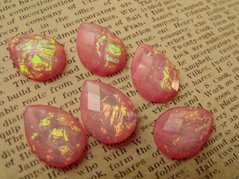 May include: Seven pink teardrop-shaped glass beads with iridescent gold flecks. The beads have a faceted design and are scattered on a brown surface.
