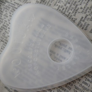 May include: A white plastic planchette with a circular hole in the centre. The planchette has the letters of the alphabet, numbers, and the words "yes", "no", and "goodbye" inscribed on it.