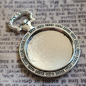 Pode incluir: Pingente prateado com uma moldura redonda e uma fivela decorativa. A moldura é projetada para segurar um cabochão redondo.