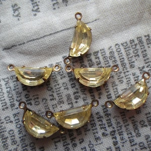 May include: Several yellow crystal pendants with a half-moon shape and small loops for hanging. The pendants have a gold-colored metal frame and are displayed on a textured, light-colored fabric.