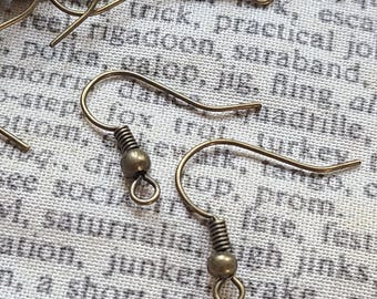 May include: Close-up of several antique brass-colored earring hooks. Each hook features a coiled spring, a small bead, and a looped end. The hooks are scattered on a patterned fabric background.