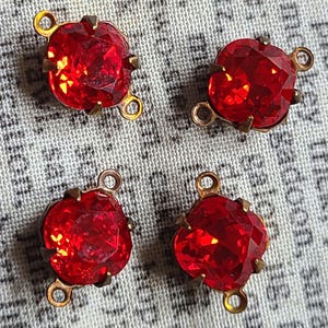 May include: Four square-shaped red faceted glass connectors with metallic prongs. Each piece features a brilliant red stone set in a brass-toned metal frame with two small circular loops on opposite sides for attachment to jewelry or craft projects.