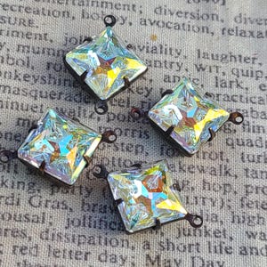 May include: Four iridescent square-cut crystal pendants with bronze-colored metal settings and loops. The crystals reflect a rainbow of colors. The pendants are displayed on a textured, printed fabric background.