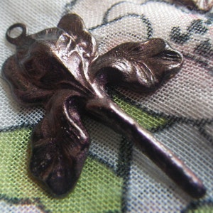 May include: A bronze-colored metal iris flower pendant with a loop at the top for hanging. The pendant has detailed petals and a stem. The flower is approximately 2 inches long.