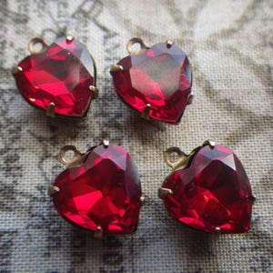 May include: Four heart-shaped red glass jewels with a faceted design. Each jewel has a brass-colored metal setting and a small loop at the top. The jewels are arranged on a patterned fabric background.