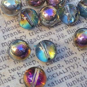 May include: A collection of spherical glass beads featuring an iridescent finish that reflects shades of purple, blue, and gold. Each bead has a distinct vertical channel or groove running through the center, creating a reflective surface.