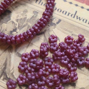 May include: Close-up of a necklace and loose beads. The necklace is a string of small, dark red beads. The loose beads are clustered together, also dark red, and flower-shaped. The background is a vintage paper.