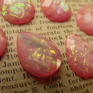 May include: A collection of pink iridescent glass teardrop shaped beads. The beads have a pearlescent finish and are scattered on a brown surface.