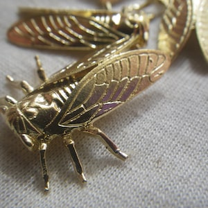 May include: Close-up of several gold-colored cicada brooches. The brooches are detailed with textured wings and legs, and are arranged on a light-colored fabric. The cicadas are in various positions, with some in focus and others blurred.