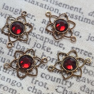 May include: Four antique brass-coloured flower-shaped connectors, each with a central, round, deep red gemstone. The connectors feature a filigree design and small loops for attachment. The background is a textured, off-white fabric.