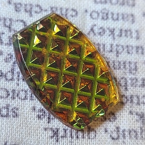 May include: A rectangular, iridescent glass bead with a textured, geometric pattern. The bead's colors shift from gold to green, with a grid of raised squares. The edges are rounded.