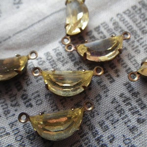 May include: Close-up of several yellow-toned, half-moon shaped crystal pendants with small loops for jewelry making. The crystals are set in a gold-colored metal. The background is a fabric with printed text.