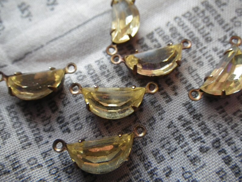 May include: Close-up of several yellow-toned, half-moon shaped crystal pendants with small loops for jewelry making. The crystals are set in a gold-colored metal. The background is a fabric with printed text.