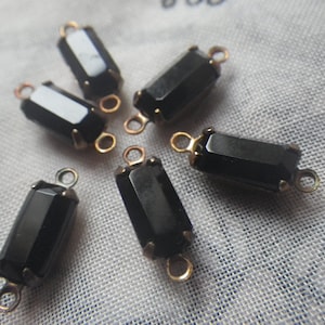 Könnte beinhalten: Sechs schwarze, rechteckige Perlen mit goldfarbenen Metallakzenten und kleinen Schlaufen zum Auffädeln. Die Perlen haben ein facettiertes Design und sind auf einem hellen Stoff angeordnet.