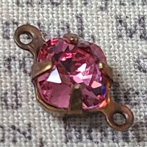 May include: A pink glass rhinestone with a gold metal setting and two loops on either side. The rhinestone is faceted and has a shiny finish.