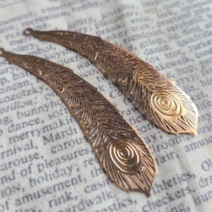 May include: Two gold-tone metal peacock feather earrings with intricate cut-out designs. The feathers have a circular eye pattern in the center.