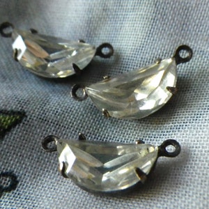 May include: Three clear, crescent-shaped crystal pendants with silver-tone metal settings and small loops for attaching to jewelry. The crystals have multiple facets, reflecting light. The pendants are displayed on a light blue fabric.