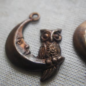 May include: A bronze charm featuring an owl perched on a crescent moon. The owl has detailed feathers and large eyes. The moon has a face with a slightly open mouth.