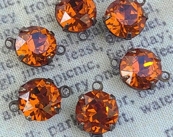 Puede incluir: Cinco piedras redondas de cristal naranja facetado en monturas de metal oscuro. Cada piedra presenta un diseño circular de talla brillante con cuatro pequeñas garras metálicas. Dos pequeñas anillas metálicas sobresalen de cada lado.
