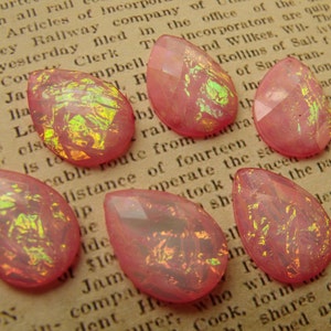 May include: Seven pink teardrop-shaped glass beads with iridescent gold and green flecks. The beads are scattered on a page of a book.