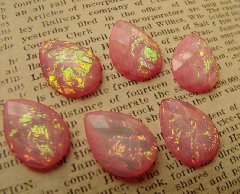 May include: Seven pink teardrop-shaped glass beads with iridescent gold and green flecks. The beads are scattered on a page of a book.