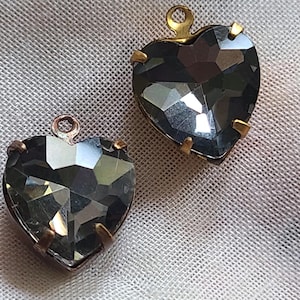 Puede incluir: Dos colgantes en forma de corazón con cristales grises facetados. Cada colgante tiene un marco de metal dorado y un pequeño lazo en la parte superior para colgar. Los colgantes se muestran sobre una superficie texturizada blanquecina.