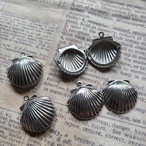 May include: Four silver-toned metal shell-shaped lockets. Three lockets are closed, showing a ribbed exterior, while one locket is open, revealing a hollow interior with matching ribbed detailing and a small circular loop at the top for attachment.