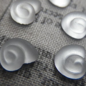 May include: Close-up of several frosted glass spiral-shaped beads. The beads are a translucent, milky white color and have a smooth, rounded appearance. They are scattered on a textured gray surface, suggesting a craft or jewelry-making theme.