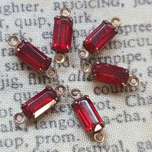 May include: Close-up of six rectangular red glass beads with gold-colored metal findings. Each bead has a faceted surface and is connected to a loop on each end. The beads are arranged in a circular pattern.