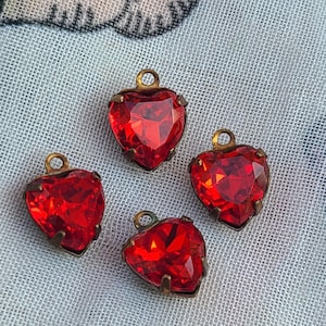 Könnte beinhalten: Vier herzförmige Anhänger mit facettierten roten Edelsteinen. Jeder Anhänger hat einen goldfarbenen Metallrahmen und eine kleine Schlaufe oben zum Befestigen an Schmuck. Die Anhänger werden auf einem hellblauen Stoff präsentiert.