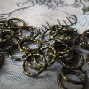 May include: A pile of gold-colored metal jump rings, each with a twisted design, on a white fabric background.