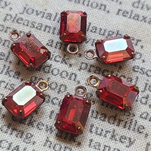 Puede incluir: Seis colgantes rectangulares de vidrio rojo facetado con engastes de metal color latón y pequeños bucles. Los colgantes están dispuestos en un patrón circular, mostrando su color vibrante y diseño intrincado. Ideal para la fabricación de joyas.