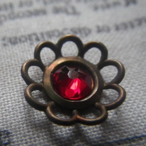 May include: A red gemstone set in a bronze flower-shaped setting. The setting has a scalloped edge.