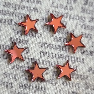 May include: Six small, star-shaped embellishments with a reflective, orange-red surface and a dark red outline. The stars are scattered on a light grey, textured fabric background.