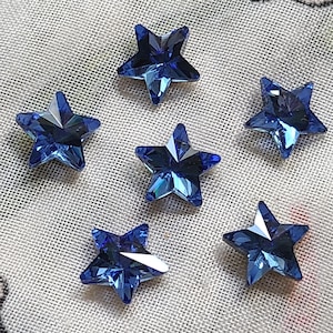 Puede incluir: Seis cuentas facetadas con forma de estrella en un color azul real intenso. Cada cuenta presenta una superficie con múltiples facetas que refleja la luz, creando bordes geométricos definidos y una apariencia brillante y centelleante en el diseño de cinco puntas.