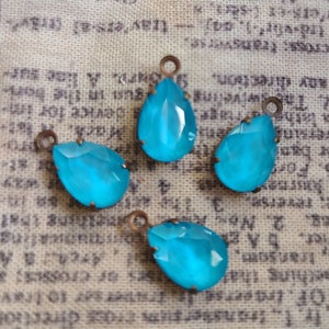 May include: Four turquoise teardrop-shaped glass charms with bronze metal loops. The charms are on a beige fabric background.