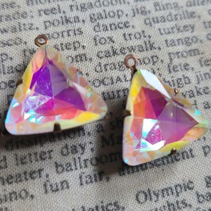 May include: Two iridescent, triangular crystal pendants with copper-colored loops. The crystals display a spectrum of colors, including pink, purple, yellow, and blue. The pendants are set in a copper-colored metal frame, showcasing a geometric design.