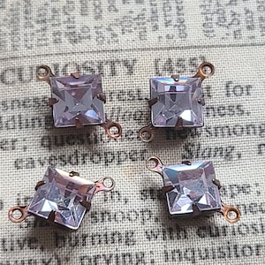 May include: Four square, light purple rhinestones with copper-colored metal loops. The rhinestones are faceted and set in a copper-colored metal frame. The background is a vintage newspaper.