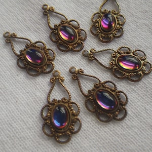 May include: Six antique brass-toned teardrop-shaped earring components. Each features an oval, iridescent glass cabochon in shades of purple, blue, and yellow, set within a decorative filigree frame. The components have a loop at the top for attachment.