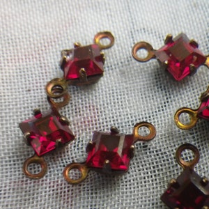 May include: A group of six red square-shaped glass rhinestones with gold metal settings. Each rhinestone has two loops on the back for attaching to a project.