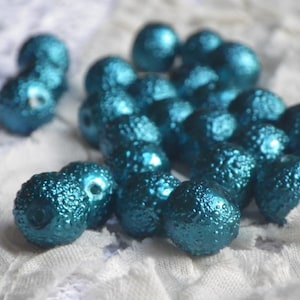 Puede incluir: Una colección de cuentas texturizadas de color turquesa. Las cuentas tienen un brillo metálico y una superficie rugosa y con bultos. Están dispersas sobre una tela blanca texturizada. Las cuentas son redondas y parecen tener un agujero para ensartar.